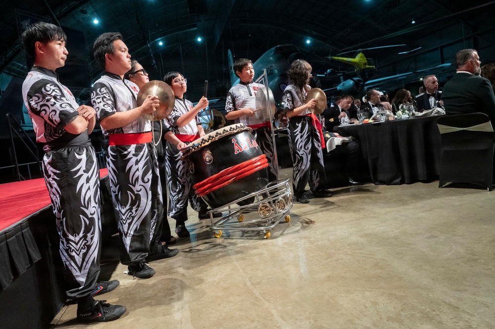 A traditional Asian drumming ensemble performs at a formal gala or banquet event held inside what appears to be an aviation museum or hangar. The performers wear matching black and white tribal-patterned uniforms with red sashes, playing large taiko-style drums and cymbals. Formally dressed guests are seated at black-draped tables in the background.