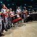 A traditional Asian drumming ensemble performs at a formal gala or banquet event held inside what appears to be an aviation museum or hangar. The performers wear matching black and white tribal-patterned uniforms with red sashes, playing large taiko-style drums and cymbals. Formally dressed guests are seated at black-draped tables in the background.
