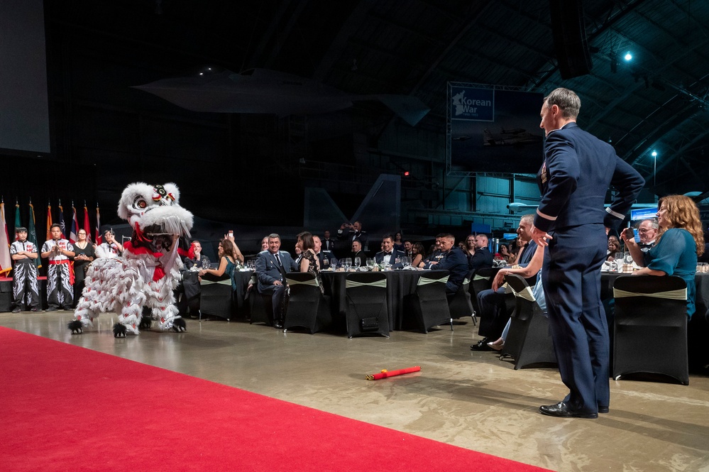 A formal military banquet event is taking place inside what appears to be an aircraft hangar, featuring a traditional Chinese/Asian lion dance performance. A uniformed military officer stands on a red carpet facing the lion dance costume, while seated guests in formal attire watch from banquet tables. Military aircraft and a 'Korean War' exhibit sign are visible in the background.