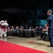 A formal military banquet event is taking place inside what appears to be an aircraft hangar, featuring a traditional Chinese/Asian lion dance performance. A uniformed military officer stands on a red carpet facing the lion dance costume, while seated guests in formal attire watch from banquet tables. Military aircraft and a 'Korean War' exhibit sign are visible in the background.