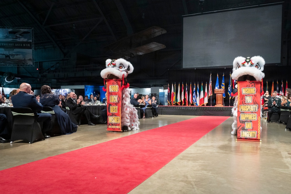 A formal banquet or military gala event featuring a traditional Chinese lion dance performance along a red carpet runway. Two white lion dance costumes are positioned on tall red decorative pillars/poles flanking the carpet, with messages of good wishes displayed. The audience, appearing to include military personnel and formally dressed guests, is seated at banquet tables on both sides.