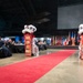 A formal banquet or military gala event featuring a traditional Chinese lion dance performance along a red carpet runway. Two white lion dance costumes are positioned on tall red decorative pillars/poles flanking the carpet, with messages of good wishes displayed. The audience, appearing to include military personnel and formally dressed guests, is seated at banquet tables on both sides.