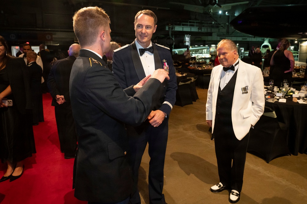 A formal military gala or ball is taking place in what appears to be a large venue, possibly an aircraft museum or hangar. Three men are engaged in conversation in the foreground: two in military dress uniforms and one in a white tuxedo jacket with military medals. The background shows additional formally dressed attendees, banquet tables, and what appears to be an aircraft suspended from the ceiling.