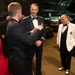 A formal military gala or ball is taking place in what appears to be a large venue, possibly an aircraft museum or hangar. Three men are engaged in conversation in the foreground: two in military dress uniforms and one in a white tuxedo jacket with military medals. The background shows additional formally dressed attendees, banquet tables, and what appears to be an aircraft suspended from the ceiling.