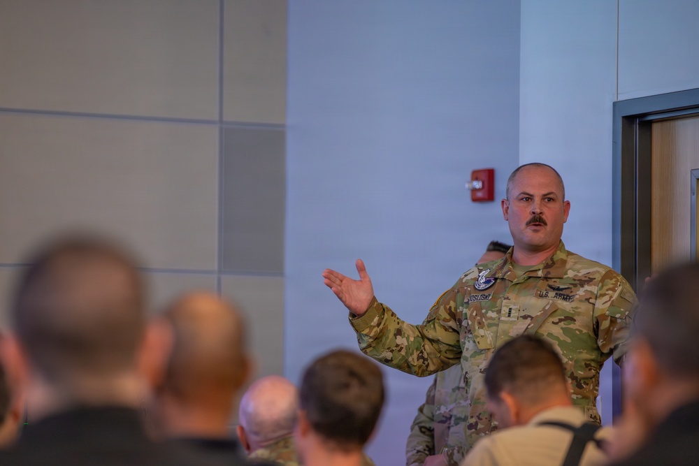 A U.S. Army soldier in OCP (Operational Camouflage Pattern) uniform is addressing a group of people in what appears to be a briefing or meeting room. The soldier is gesturing with one hand while speaking to an audience seated in front of him. The setting appears to be a modern military or government facility.