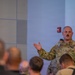 A U.S. Army soldier in OCP (Operational Camouflage Pattern) uniform is addressing a group of people in what appears to be a briefing or meeting room. The soldier is gesturing with one hand while speaking to an audience seated in front of him. The setting appears to be a modern military or government facility.