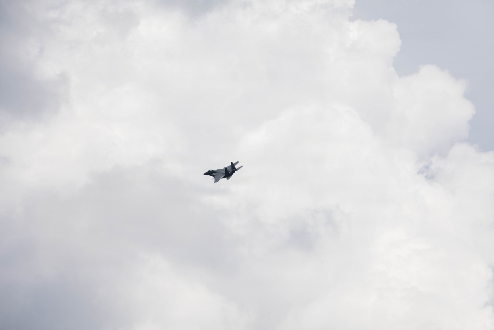 A single military jet aircraft is captured in flight against an overcast, cloudy sky. The aircraft appears to be banking or maneuvering, visible near the center of the frame. The image has a muted, grey tone due to the heavy cloud cover.