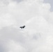 A single military jet aircraft is captured in flight against an overcast, cloudy sky. The aircraft appears to be banking or maneuvering, visible near the center of the frame. The image has a muted, grey tone due to the heavy cloud cover.