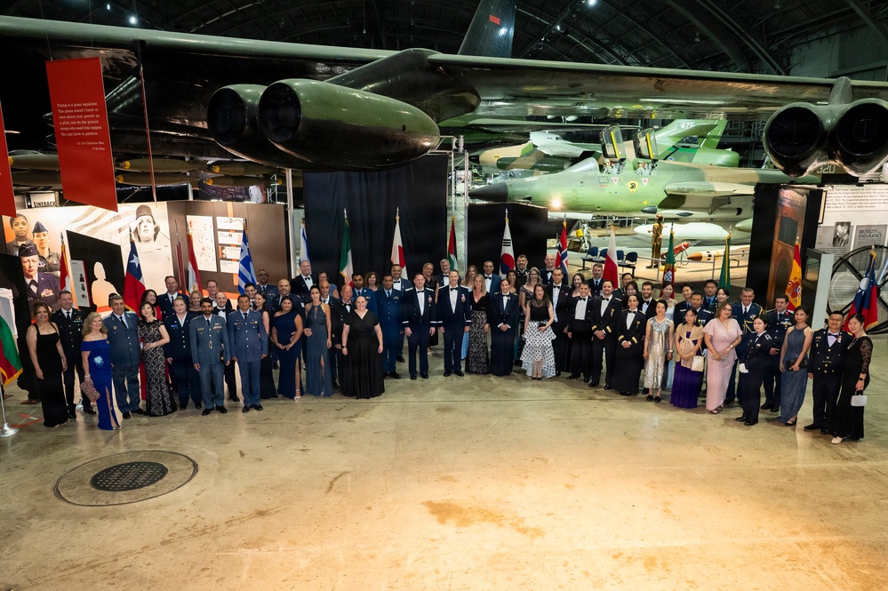 A large formal group photograph taken inside what appears to be an aviation museum or air force hangar, with a large military aircraft suspended overhead. The group consists of approximately 60-70 individuals dressed in formal attire including military dress uniforms and evening gowns, suggesting an official international military ceremony or gala. Multiple national flags are visible in the background, indicating an international or multinational military event.