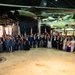 A large formal group photograph taken inside what appears to be an aviation museum or air force hangar, with a large military aircraft suspended overhead. The group consists of approximately 60-70 individuals dressed in formal attire including military dress uniforms and evening gowns, suggesting an official international military ceremony or gala. Multiple national flags are visible in the background, indicating an international or multinational military event.