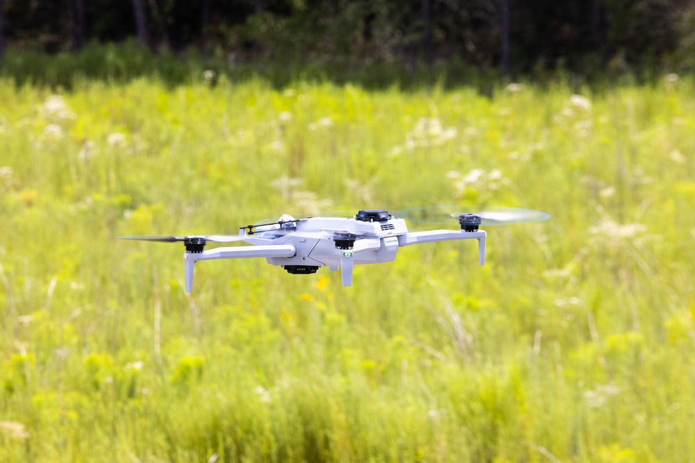 A white consumer quadcopter drone is captured mid-flight over a lush green meadow or field. The drone is in sharp focus against a softly blurred background of tall grasses, wildflowers, and trees. The image appears to be taken during daytime in warm, sunny conditions.