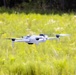 A white consumer quadcopter drone is captured mid-flight over a lush green meadow or field. The drone is in sharp focus against a softly blurred background of tall grasses, wildflowers, and trees. The image appears to be taken during daytime in warm, sunny conditions.