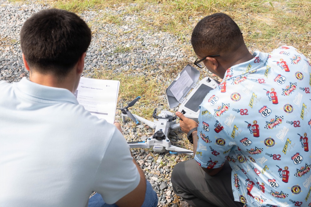 Two individuals are crouched on a gravel surface examining a small consumer drone, likely a DJI model. One person holds a document that appears to be a flight card or checklist, while the other operates a remote controller with an attached screen. They appear to be conducting a pre-flight inspection or setup procedure.