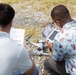 Two individuals are crouched on a gravel surface examining a small consumer drone, likely a DJI model. One person holds a document that appears to be a flight card or checklist, while the other operates a remote controller with an attached screen. They appear to be conducting a pre-flight inspection or setup procedure.
