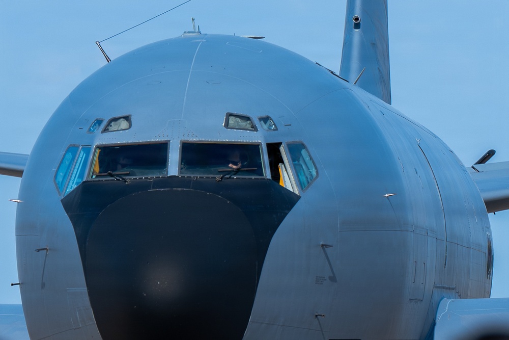 A close-up frontal view of a large military aircraft, likely a Boeing KC-135 Stratotanker or similar tanker/transport aircraft, painted in standard military gray. The image captures the nose section and cockpit in sharp detail, with a crew member visible through the cockpit windows. The aircraft appears to be on the ground or taxiing, photographed against a clear blue sky.