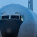 A close-up frontal view of a large military aircraft, likely a Boeing KC-135 Stratotanker or similar tanker/transport aircraft, painted in standard military gray. The image captures the nose section and cockpit in sharp detail, with a crew member visible through the cockpit windows. The aircraft appears to be on the ground or taxiing, photographed against a clear blue sky.