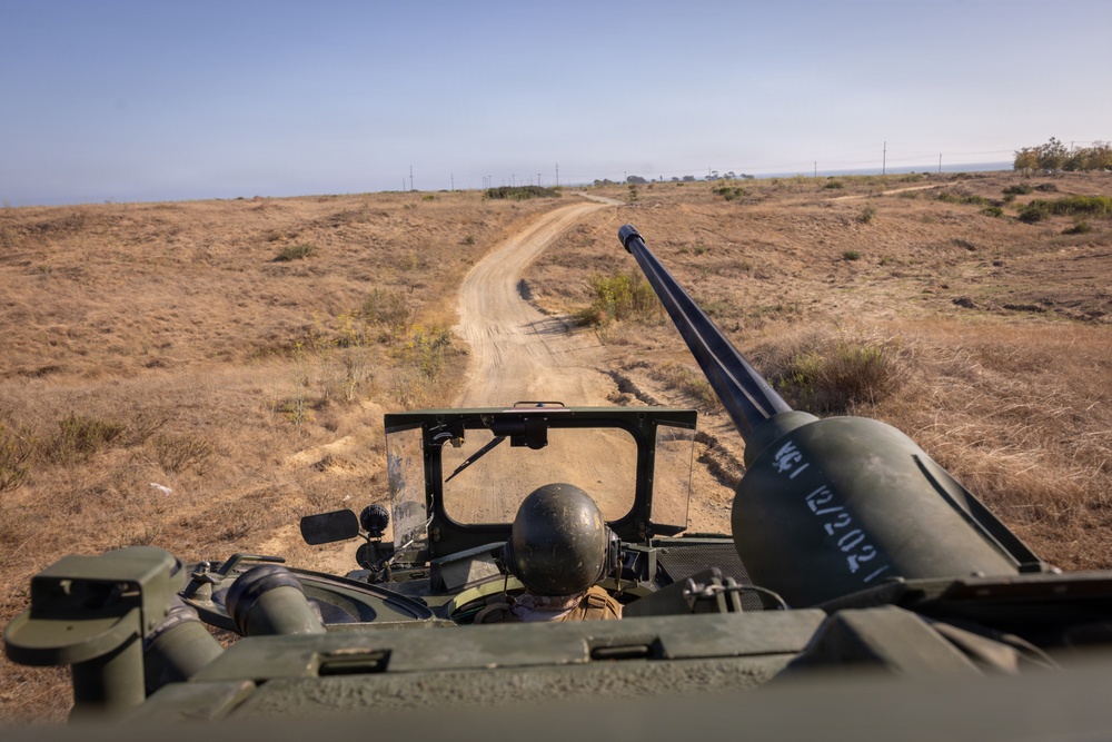 The image is taken from the top of a military armored vehicle traveling along a dirt road through dry, open terrain. A soldier wearing a helmet is visible in the driver's hatch below, and a weapon system or recoilless rifle is mounted prominently in the foreground. The landscape appears arid with sparse vegetation, utility poles visible in the distance, and what appears to be a coastline or body of water on the horizon.