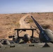 The image is taken from the top of a military armored vehicle traveling along a dirt road through dry, open terrain. A soldier wearing a helmet is visible in the driver's hatch below, and a weapon system or recoilless rifle is mounted prominently in the foreground. The landscape appears arid with sparse vegetation, utility poles visible in the distance, and what appears to be a coastline or body of water on the horizon.