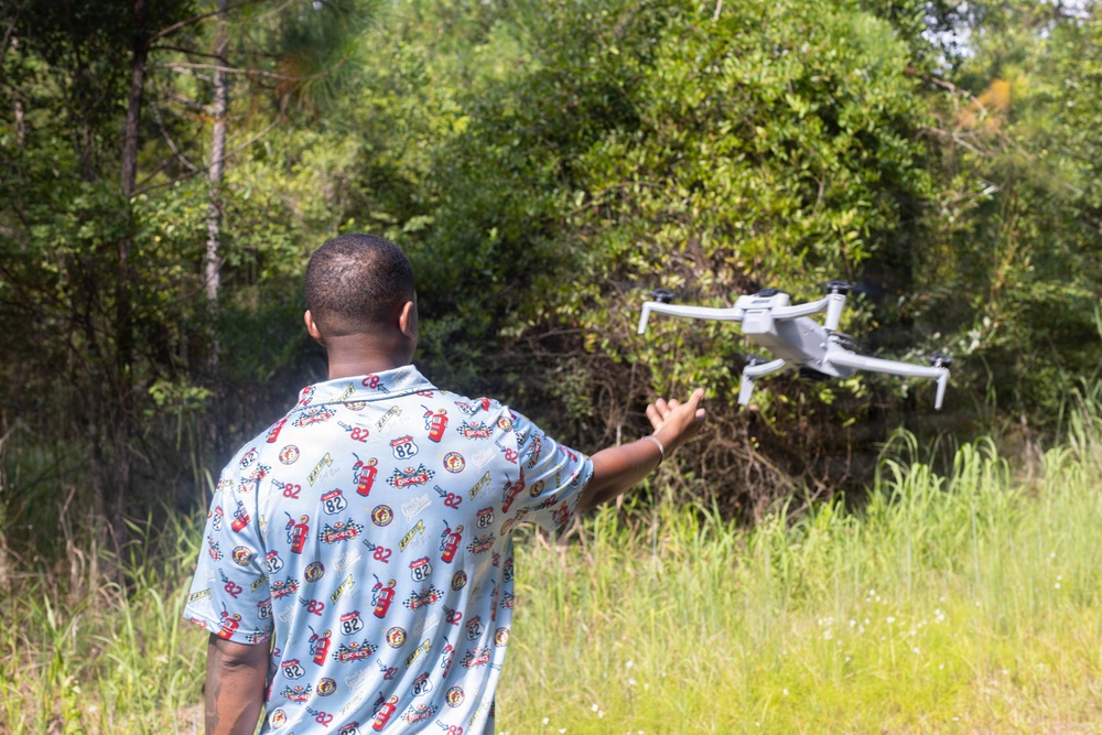 A man is seen from behind launching or catching a white quadcopter drone in an outdoor, vegetated setting. He is wearing a light blue patterned shirt with racing and fire extinguisher graphics, and his right arm is extended toward the drone. The background consists of dense green trees and tall grass, suggesting a rural or semi-wild environment.