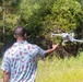 A man is seen from behind launching or catching a white quadcopter drone in an outdoor, vegetated setting. He is wearing a light blue patterned shirt with racing and fire extinguisher graphics, and his right arm is extended toward the drone. The background consists of dense green trees and tall grass, suggesting a rural or semi-wild environment.