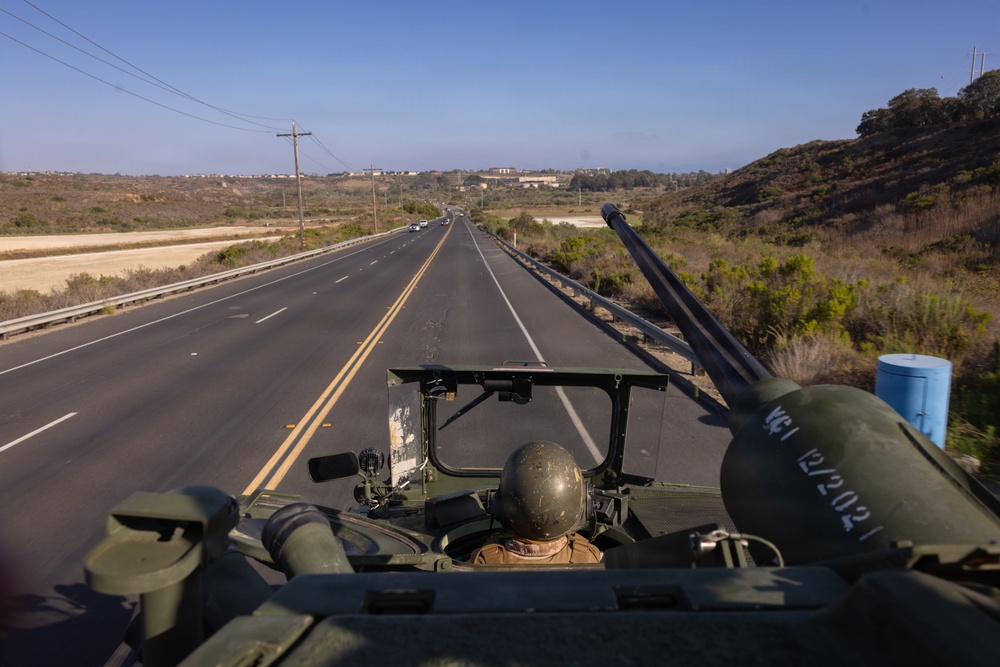 A military vehicle is traveling along a two-lane highway through a semi-arid coastal landscape, viewed from a rear elevated perspective. A soldier wearing a helmet is visible in the vehicle's open hatch, with what appears to be a weapon system or antenna mounted nearby. The surrounding terrain features dry scrubland and low hills, consistent with Southern California geography.