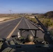 A military vehicle is traveling along a two-lane highway through a semi-arid coastal landscape, viewed from a rear elevated perspective. A soldier wearing a helmet is visible in the vehicle's open hatch, with what appears to be a weapon system or antenna mounted nearby. The surrounding terrain features dry scrubland and low hills, consistent with Southern California geography.