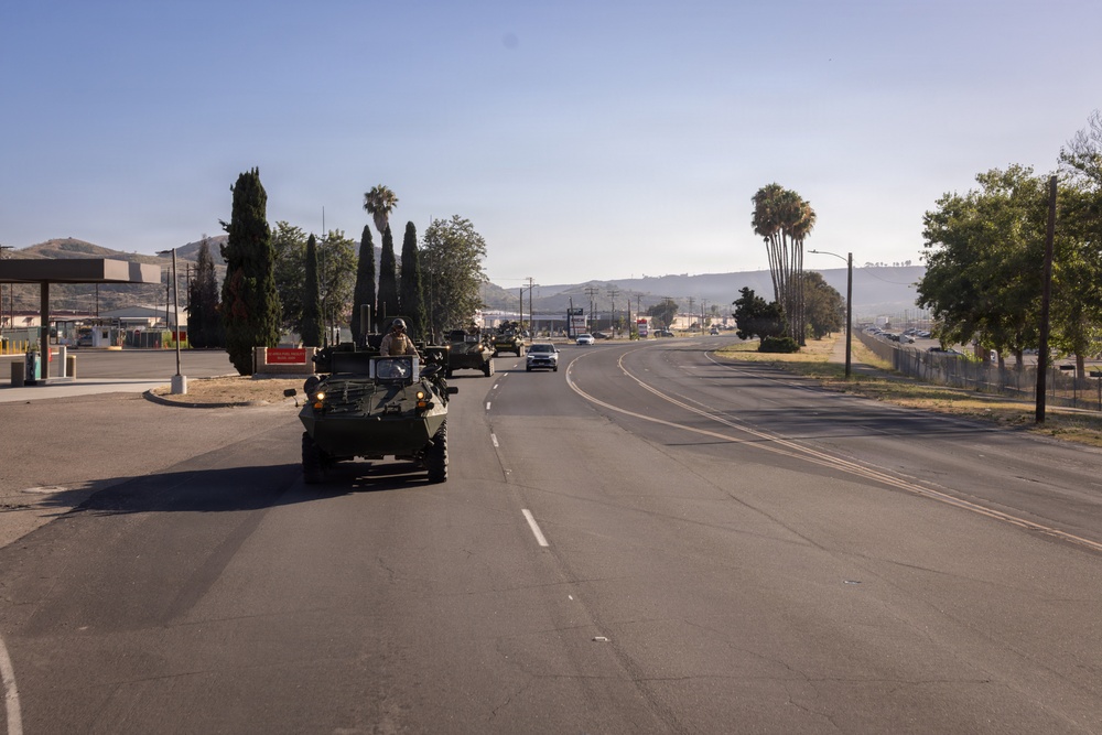 A convoy of military armored vehicles is traveling along a paved road in what appears to be a semi-arid region, likely a U.S. military installation or adjacent public road. At least two light armored vehicles (LAVs) are visible in the foreground, with additional military vehicles further down the road. The setting features palm trees, dry hills, and civilian infrastructure consistent with Southern California.