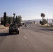 A convoy of military armored vehicles is traveling along a paved road in what appears to be a semi-arid region, likely a U.S. military installation or adjacent public road. At least two light armored vehicles (LAVs) are visible in the foreground, with additional military vehicles further down the road. The setting features palm trees, dry hills, and civilian infrastructure consistent with Southern California.