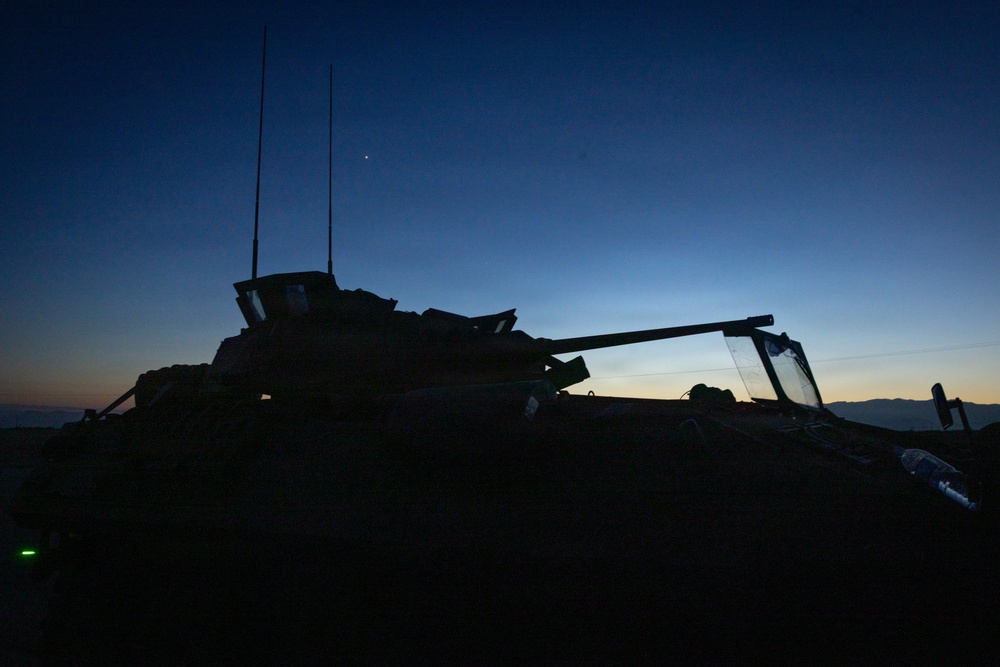 A military armored vehicle is photographed in silhouette against a twilight sky at dusk or dawn. The vehicle's turret, gun barrel, and radio antennas are clearly outlined against the gradient blue-orange horizon. A small green indicator light is visible at the lower left, and distant mountains are faintly visible on the horizon.