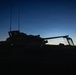A military armored vehicle is photographed in silhouette against a twilight sky at dusk or dawn. The vehicle's turret, gun barrel, and radio antennas are clearly outlined against the gradient blue-orange horizon. A small green indicator light is visible at the lower left, and distant mountains are faintly visible on the horizon.