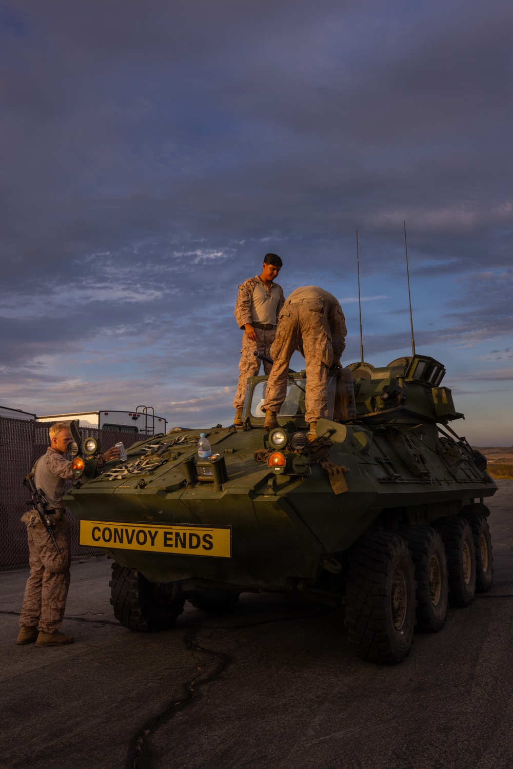 Three military personnel in desert camouflage uniforms are conducting maintenance or inspection on a green eight-wheeled Light Armored Vehicle (LAV) at dusk or dawn. Two personnel are positioned on top of the vehicle while a third stands at the front. The vehicle displays a yellow sign reading 'CONVOY ENDS,' indicating it is the last vehicle in a convoy formation.