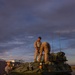 Three military personnel in desert camouflage uniforms are conducting maintenance or inspection on a green eight-wheeled Light Armored Vehicle (LAV) at dusk or dawn. Two personnel are positioned on top of the vehicle while a third stands at the front. The vehicle displays a yellow sign reading 'CONVOY ENDS,' indicating it is the last vehicle in a convoy formation.