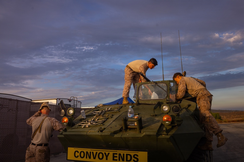 Military personnel, appearing to be U.S. Marines based on their camouflage uniforms, are working on or around a green armored military vehicle marked 'CONVOY ENDS' on its front. The scene takes place at dusk or dawn, with a dramatic cloudy sky in the background. The vehicle appears to be an LAV (Light Armored Vehicle) or similar wheeled armored personnel carrier.