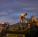 Military personnel, appearing to be U.S. Marines based on their camouflage uniforms, are working on or around a green armored military vehicle marked 'CONVOY ENDS' on its front. The scene takes place at dusk or dawn, with a dramatic cloudy sky in the background. The vehicle appears to be an LAV (Light Armored Vehicle) or similar wheeled armored personnel carrier.