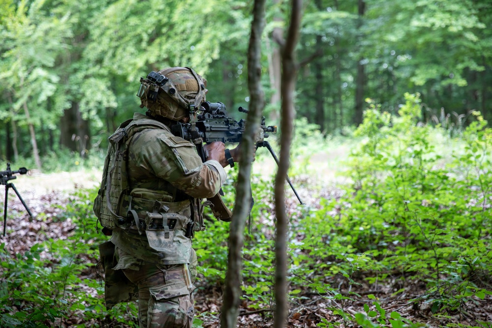A fully equipped soldier in multicam camouflage is aiming a rifle with an optical scope in a dense woodland environment. The soldier is wearing a tactical helmet with mounted accessories, a plate carrier vest, and a full combat load. A second weapon system on a bipod is partially visible in the background left.