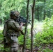 A fully equipped soldier in multicam camouflage is aiming a rifle with an optical scope in a dense woodland environment. The soldier is wearing a tactical helmet with mounted accessories, a plate carrier vest, and a full combat load. A second weapon system on a bipod is partially visible in the background left.