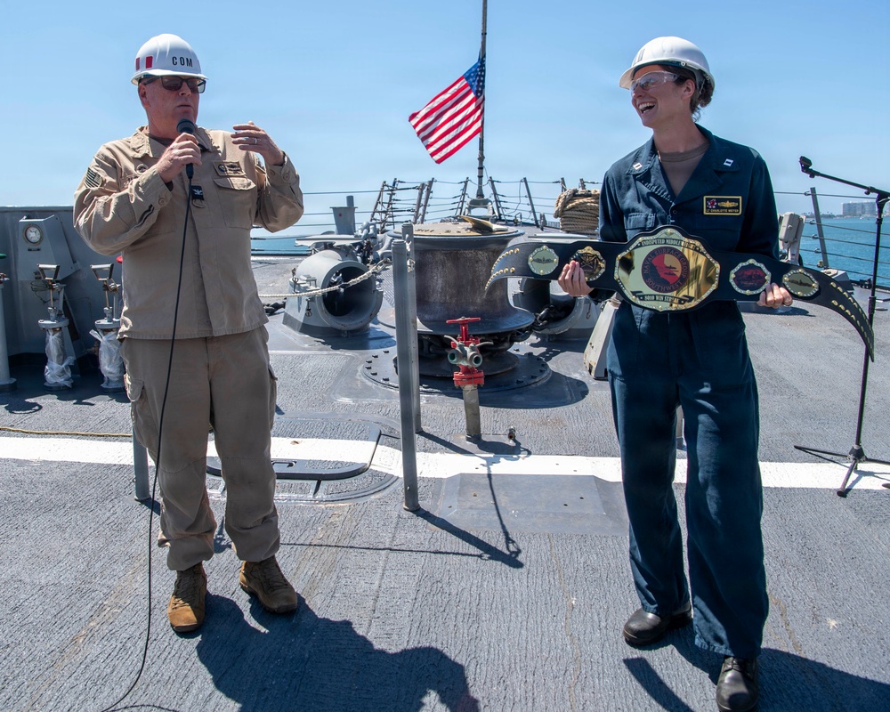 Two military personnel are standing on the deck of a naval vessel during what appears to be an award or recognition ceremony. One individual in tan/khaki uniform holds a microphone and wears a hard hat labeled 'COM', while the other in dark coveralls holds a large championship-style belt and is smiling. An American flag is visible in the background.
