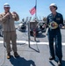 Two military personnel are standing on the deck of a naval vessel during what appears to be an award or recognition ceremony. One individual in tan/khaki uniform holds a microphone and wears a hard hat labeled 'COM', while the other in dark coveralls holds a large championship-style belt and is smiling. An American flag is visible in the background.
