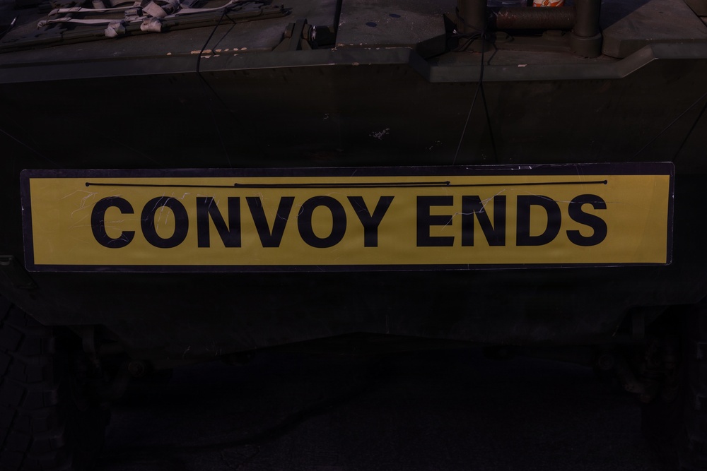 The image shows a yellow rectangular sign with bold black text reading 'CONVOY ENDS', mounted on what appears to be a dark-colored military vehicle. The sign features a black border and two horizontal black lines above the text. The setting appears to be an indoor or low-light environment, likely a garage or depot.