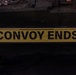The image shows a yellow rectangular sign with bold black text reading 'CONVOY ENDS', mounted on what appears to be a dark-colored military vehicle. The sign features a black border and two horizontal black lines above the text. The setting appears to be an indoor or low-light environment, likely a garage or depot.