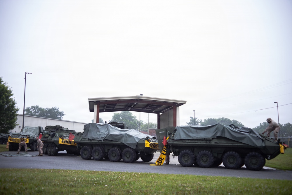 The image shows multiple large military wheeled armored vehicles, consistent with Stryker-type APCs, parked in a convoy formation on a paved area near a covered structure. The vehicles are covered with olive-drab tarps and display yellow 'WIDE LOAD' warning signs, indicating they have recently been or are being transported on public roads. Military personnel in camouflage uniforms are visible working around the vehicles.