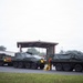 The image shows multiple large military wheeled armored vehicles, consistent with Stryker-type APCs, parked in a convoy formation on a paved area near a covered structure. The vehicles are covered with olive-drab tarps and display yellow 'WIDE LOAD' warning signs, indicating they have recently been or are being transported on public roads. Military personnel in camouflage uniforms are visible working around the vehicles.