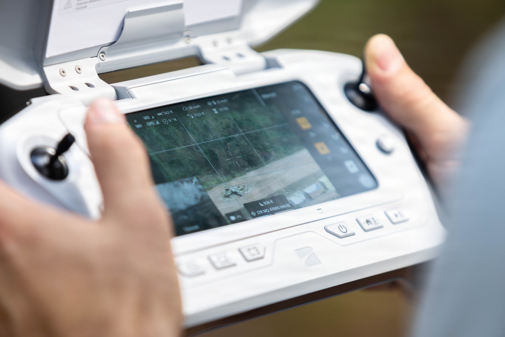 A person is holding a white drone remote controller with an integrated screen displaying a live camera feed from an aerial drone. The screen shows the drone hovering over a wooded area with telemetry data overlaid, including altitude, GPS coordinates, and speed readings. The controller appears to be a DJI-style smart controller with built-in display and physical joysticks.