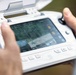 A person is holding a white drone remote controller with an integrated screen displaying a live camera feed from an aerial drone. The screen shows the drone hovering over a wooded area with telemetry data overlaid, including altitude, GPS coordinates, and speed readings. The controller appears to be a DJI-style smart controller with built-in display and physical joysticks.