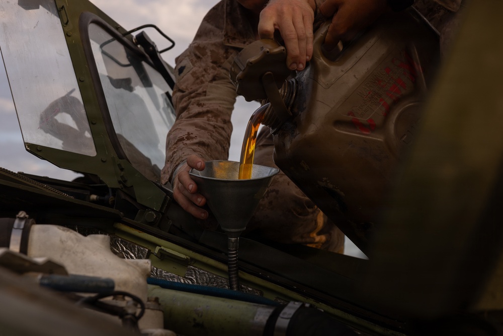 A person wearing desert camouflage military uniform is performing vehicle maintenance by pouring a golden-amber fluid, likely engine oil, through a funnel into a military vehicle's engine compartment. The vehicle hood is open, revealing engine components, and a large military-style jerry can or fluid container is being used as the source. The setting appears to be an outdoor field environment.