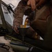 A person wearing desert camouflage military uniform is performing vehicle maintenance by pouring a golden-amber fluid, likely engine oil, through a funnel into a military vehicle's engine compartment. The vehicle hood is open, revealing engine components, and a large military-style jerry can or fluid container is being used as the source. The setting appears to be an outdoor field environment.
