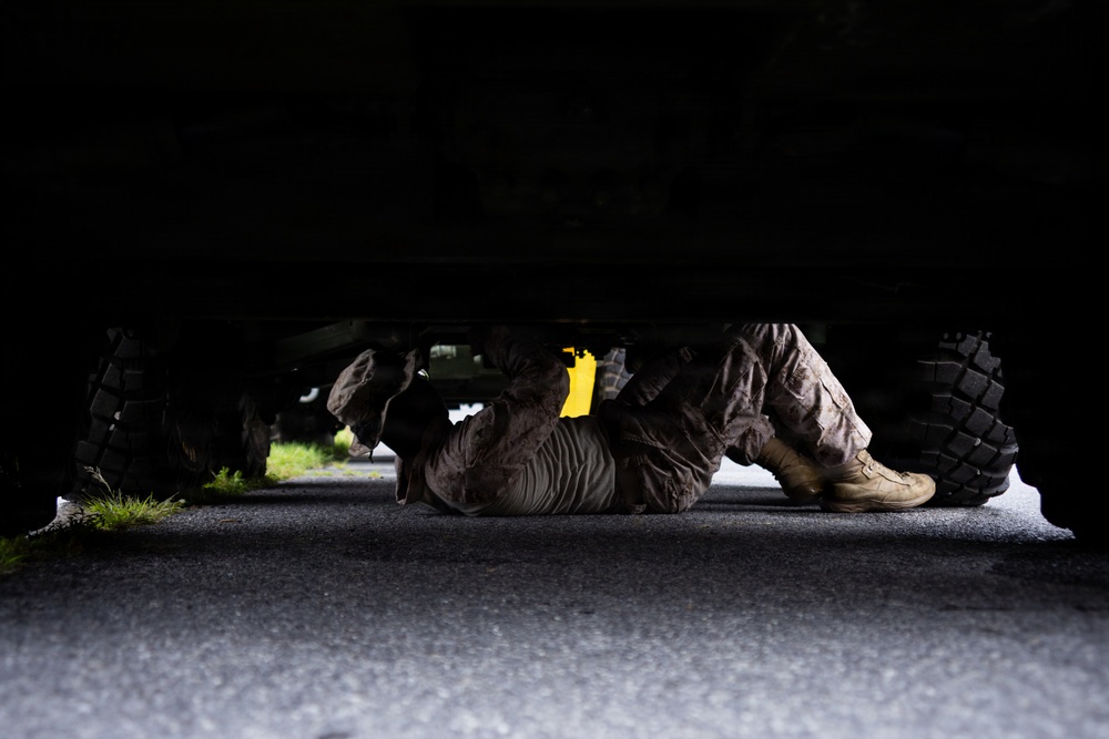 A person wearing military-style camouflage clothing and tan combat boots is lying on their back on a paved surface underneath a large vehicle. The individual appears to be performing maintenance or inspection work on the underside of the vehicle. A yellow component is visible beneath the vehicle chassis.