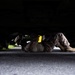 A person wearing military-style camouflage clothing and tan combat boots is lying on their back on a paved surface underneath a large vehicle. The individual appears to be performing maintenance or inspection work on the underside of the vehicle. A yellow component is visible beneath the vehicle chassis.