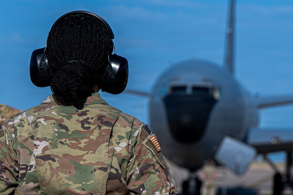 A military service member wearing OCP (Operational Camouflage Pattern) uniform and hearing protection stands on a flight line facing a large military aircraft. The individual has braided hair styled in a bun and is viewed from behind. The scene appears to be an active airfield operation with the aircraft positioned directly ahead.