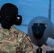 A military service member wearing OCP (Operational Camouflage Pattern) uniform and hearing protection stands on a flight line facing a large military aircraft. The individual has braided hair styled in a bun and is viewed from behind. The scene appears to be an active airfield operation with the aircraft positioned directly ahead.